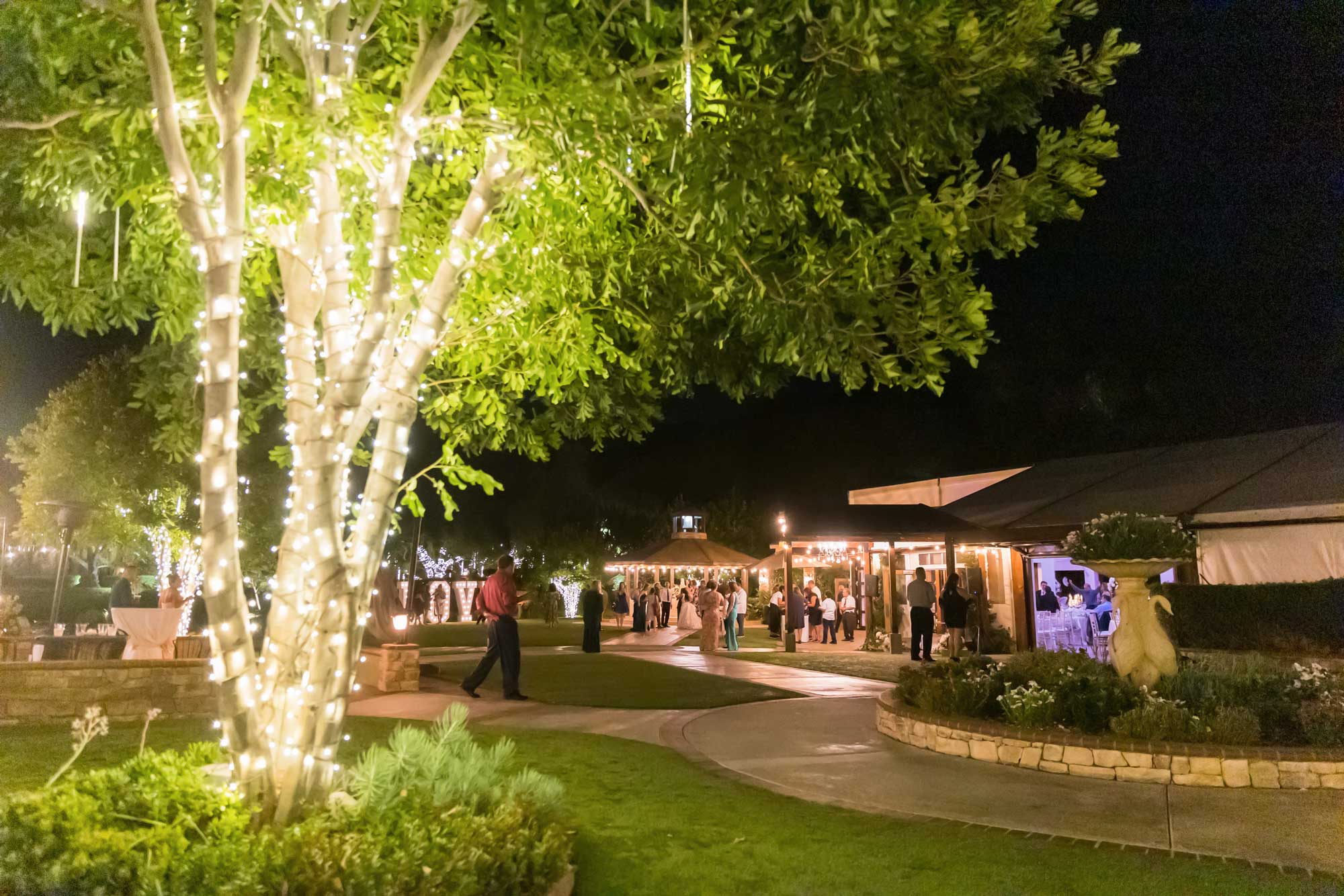 property 466 3271 Azie Christopher Outdoor nighttime event with guests mingling near a tent; trees and pathways are decorated with string lights, creating a festive atmosphere.