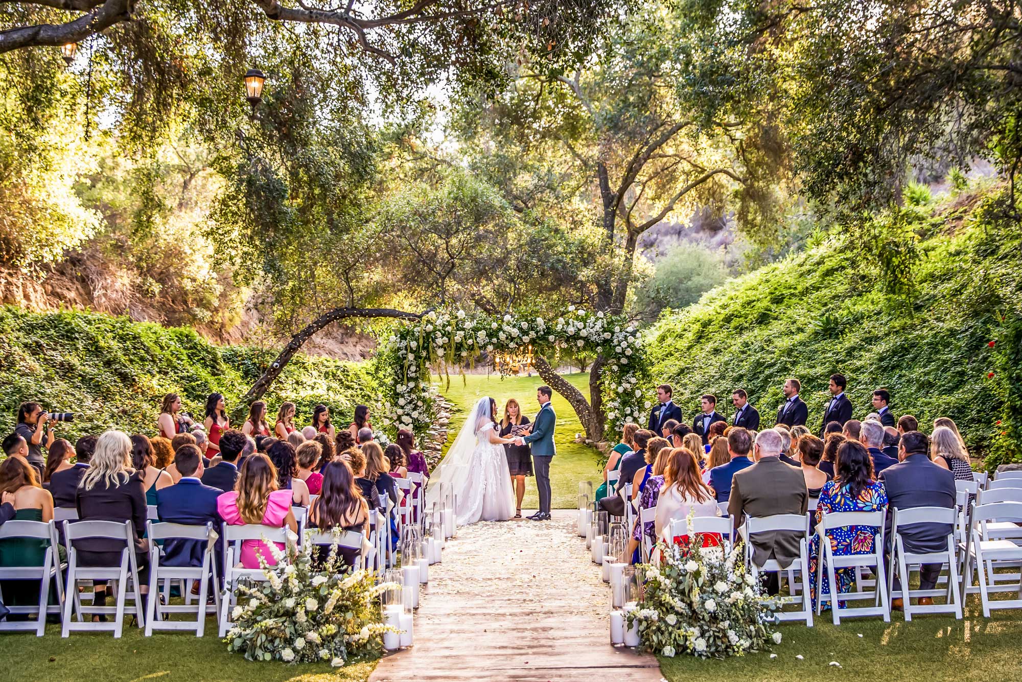 los willows ceremony 554 2298 Shay Dan Outdoor wedding ceremony with a large group of seated guests, a bride and groom standing at the altar under a floral arch, surrounded by greenery and trees.