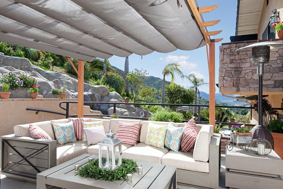 villa terrace 19 9325189 106348 bpo 106348 119 Outdoor patio -1163 features a cushioned sectional sofa, striped and patterned pillows, coffee table with decorative lantern, potted plants, sunshade, and a scenic view of hills in the background.