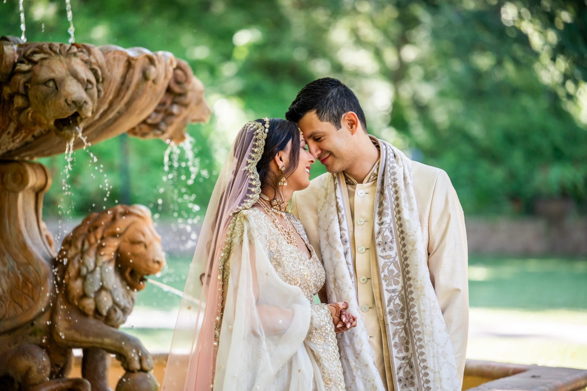 Indian1 compressed A couple in traditional South Asian wedding attire stands close together by a fountain with lion carvings, surrounded by greenery.