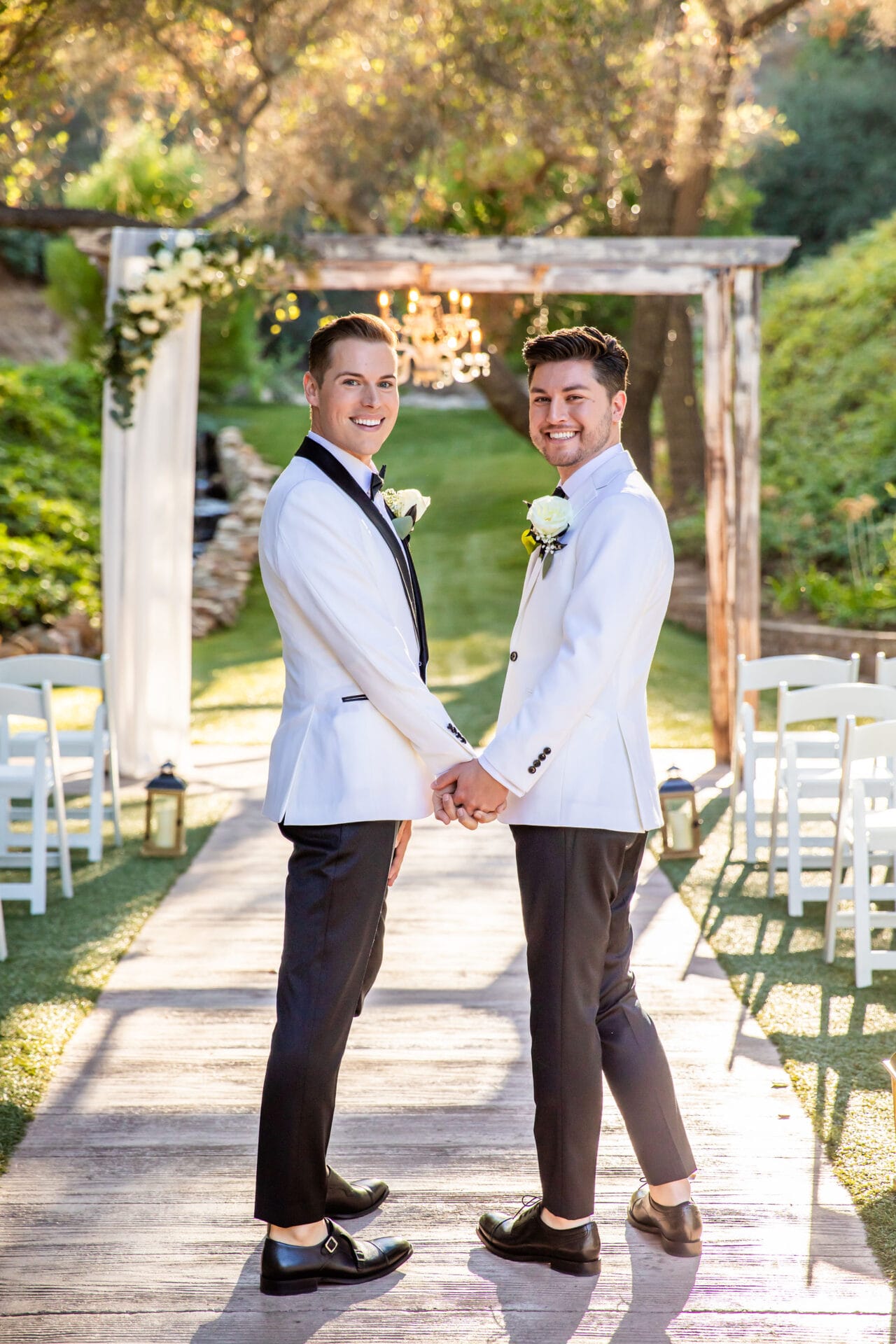2428Scott Tyler Two men in white tuxedos hold hands and smile at the camera while standing on an outdoor wedding aisle decorated with white chairs and greenery.