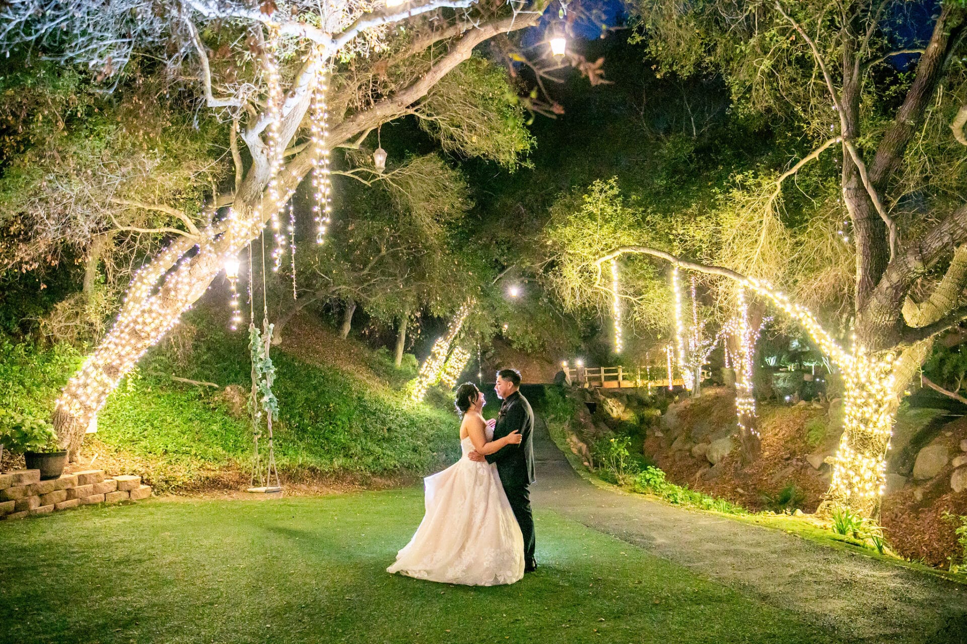 3251Gabby Aquiles A bride and groom stand together on a garden path at night, surrounded by trees decorated with string lights.
