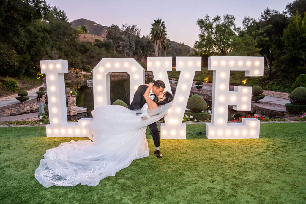 A couple in wedding attire poses in front of large, illuminated "LOVE" letters on a grassy outdoor area with trees and mountains in the background.