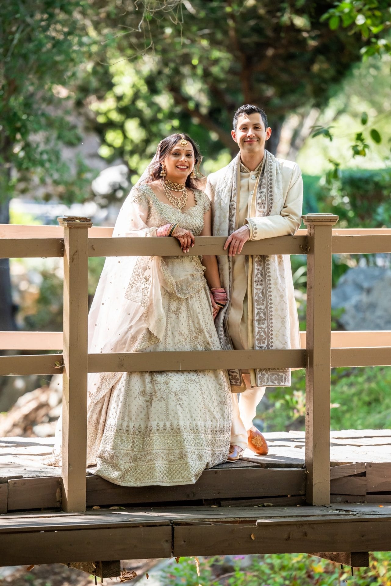 Indian Fusion wedding A couple dressed in traditional Indian wedding attire stands together on a wooden bridge in a garden setting.